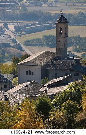 Stock Image - Aerial view of village stone church with bell tower, slate roofs, and autumn colours of trees, with plain below of planted fields, Lombardy, Italy. Fotosearch