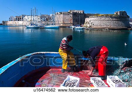 Brightly Dressed Fishermen Emptying Nets On Old Wooden Fishing Boat In Harbour With Sailboats Moored Beneath Medieval Stone Walls Of Gallipoli Sunny Spring Morning With Blue Sky Salento Puglia Southern Italy Stock
