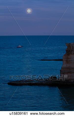 Stock Image - Cliffs and rocky coastline of Salento at twilight, with full moon reflection on sea, carabinieri motor boat in distance, Puglia, Southern Italy. Fotosearch