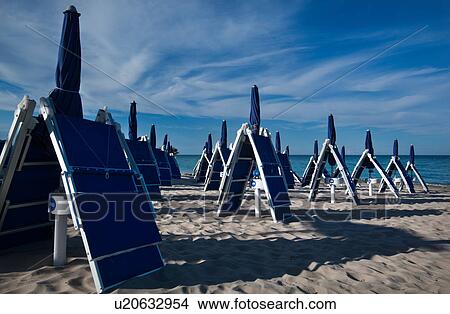 Picture - Close up of rows of folded blue beach chairs and umbrellas casting long shadows across fine sandy beach, blue calm sea and dramatic clouds in sunny blue sky, Salento, Puglia, Southern Italy. Fotosearch