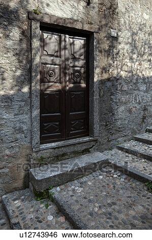 Stock Photograph - Closeup of brown wooden carved door set in medieval stone house, with pebbled steps in outside alley of village Northern Italy. Fotosearch