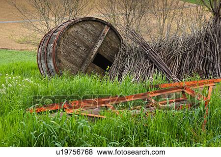 Stock Photo - Closeup of old red metal farming implements and ancient wooden barrel in grass, with stack of kindling ploughed fields in background, Umbria, Italy. Fotosearch