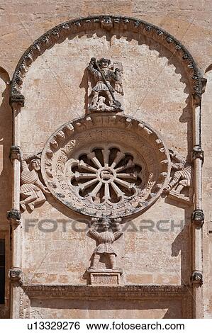Stock Photograph - Detail of Romanesque facade of church of San Domenico, with typical stone carved scroll work and statues around rose window, sunny spring afternoon, Matera, Puglia, Southern Italy. Fotosearch