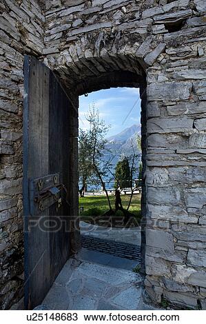 Stock Image - Doorway, Castello di Vezio, Lake Como, Northern Italy, with wooden door and ancient rusty bolt, showing view of gardens and mountain slopes beyond, sunlit morning in autumn.. Fotosearch