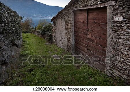 Picture - Doorway of cantina, with old double wooden plank doors in medieval stone building, with stone wall opposite flanking village pathway, view of lake and mountains beyond in late afternoon, Lake Como, Northern Italy. Fotosearch
