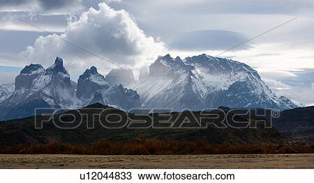 Stock Image - Early morning panorama view of snow covered Los Cuernos, and mountain range, Torres del Paine, with dramatic clouds, beech trees in full autumn colours, Southern Chile, Patagonia. Fotosearch