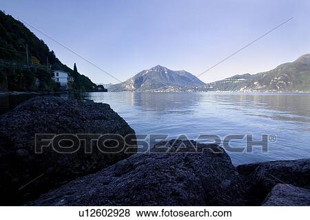 Stock Photo - Early morning view of Lake Como from rocky inlet shore, calm blue water, with cloudless blue sky, and mountains reflected in water, Lake Como, Northern Italy. Fotosearch