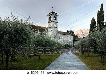 Stock Image - Exterior view of apse, bell tower and side of Abbey of Piona, Lake Como, Northern Italy from churchyard and gardens, with pathway leading to Abbey, in early morning pale sunlight, with blue sky. Fotosearch