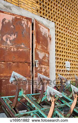 Stock Image - Exterior view of old barn with rusty iron prongs of green harrow in foreground, old red metal doors and pierced cooling detail yellow walls in background, Umbria, Italy. Fotosearch