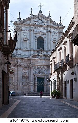 Stock Image - Facade of church of Sant'Irene, exuberant Baroque ornamentation of facade, street with single female figure in distance, sunny spring morning, Lecce, Salento, Puglia, Southern Italy. Fotosearch