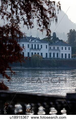 Stock Photo - Facade of Renaissance villa, with white stucco facade and arched shuttered windows across calm water of Lake Como, flanked by Japanese maple tree, on hazy sunlit autumn afternoon, Northern Italy.. Fotosearch
