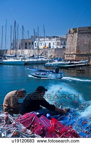 Stock Image - Fishermen mending colourful nets in harbour with sailboats moored beneath medieval stone walls of Gallipoli, sunny spring morning with blue sky, Salento, Puglia, Southern Italy. Fotosearch