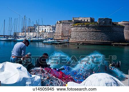 Stock Photography - Fishermen mending colourful nets in harbour with sailboats moored beneath medieval stone walls of Gallipoli, sunny spring morning with blue sky, Salento, Puglia, Southern Italy. Fotosearch
