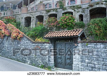 Front detail of large old house in Northern italy with stone facade, arches filled with flowers, extended balcony, with tile-covered entranceway, stone wall, and modern metal cast door Northern italy View Large Photo Image Stock Photo - Front detail of large old house in Northern italy with stone facade, arches filled with flowers, extended balcony, with tile-covered entranceway, stone wall, and modern metal cast door Northern italy. Fotosearch