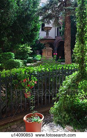 Stock Image - Internal courtyard of Renaissance palazzo with shrubs and trees beyond wrought iron railing, and central fountain, Bologna Italy. Fotosearch