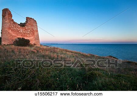 Stock Photo - Medieval defense tower overlooking coastline, calm sea, at sunset with pink light on stone tower, and pink clouds, Gallipoli town on horizon, Salento, Puglia, Southern Italy. Fotosearch