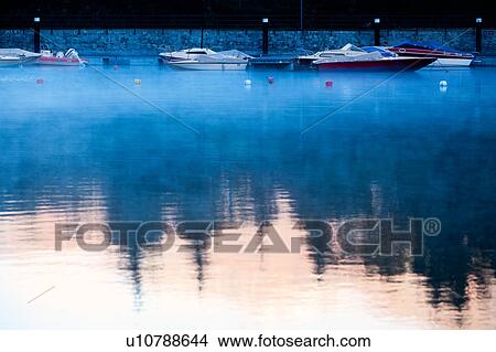 Picture - Motorboats moored at Lake Como,, golden light of sunrise and pine trees reflected in water with swirling mists, Lombardy, Northern Italy.. Fotosearch