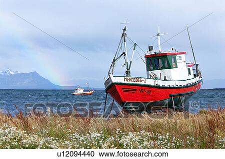 Stock Image - Old red wooden fishing boat beached with fishing boat moored beyond in choppy blue waters, rainbow and mountainsin background, Puerto Natales, Southern Chile, Patagonia. Fotosearch