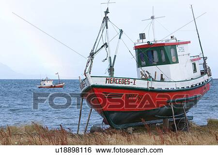 Stock Photograph - Old red wooden fishing boat beached with fishing boat moored beyond in choppy blue waters, rainbow and mountainsin background, Puerto Natales, Southern Chile, Patagonia. Fotosearch