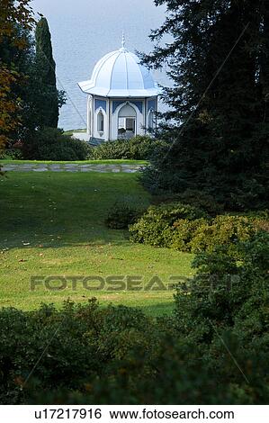 Stock Photograph - Ornamental gardens of Villa Melzi along lake shore, flanked by cypress trees leading to octagonal tempio with blue dome and arched windows and doors, sunlit afternoon, Lake Como, Northern Italy. Fotosearch
