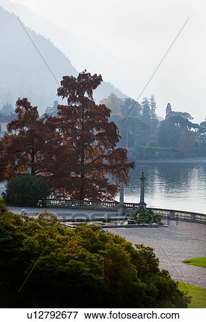 Ornamental gardens of Villa Melzi with path along lake shore, flanked by trees and bushes with autumn foliage, stone carved balustrade with stone columns, on sunlit hazy autumn afternoon, Lake Como, Northern Italy View Large Photo Image Stock Photo - Ornamental gardens of Villa Melzi with path along lake shore, flanked by trees and bushes with autumn foliage, stone carved balustrade with stone columns, on sunlit hazy autumn afternoon, Lake Como, Northern Italy. Fotosearch
