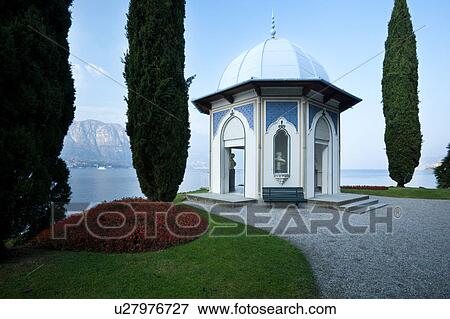 Ornamental gardens of Villa Melzi with path along lake shore, flanked by cypress trees leading to octagonal tempio with blue dome and arched windows and doors, sunlit afternoon, Lake Como, Northern Italy View Large Photo Image Stock Photo - Ornamental gardens of Villa Melzi with path along lake shore, flanked by cypress trees leading to octagonal tempio with blue dome and arched windows and doors, sunlit afternoon, Lake Como, Northern Italy. Fotosearch