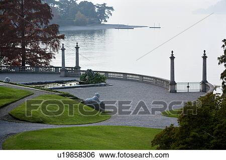 Ornamental gardens of Villa Melzi with path along lake shore, flanked by trees and bushes with autumn foliage, grassy slopes, pond with waterlilies, stone carved balustrade with stone columns, and lion statues, on sunlit hazy autumn afternoon, Lake Como, View Large Photo Image Stock Photograph - Ornamental gardens of Villa Melzi with path along lake shore, flanked by trees and bushes with autumn foliage, grassy slopes, pond with waterlilies, stone carved balustrade with stone columns, and lion statues, on sunlit hazy autumn afternoon, Lake Como, . Fotosearch