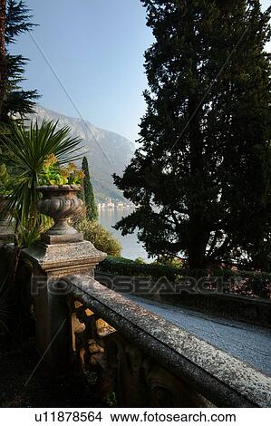 Picture - Ornamental gardens of Villa Monastero with carved stone wall, and terracotta pots on columns, with view overlooking Lake Como and mountain range beyond, autumn afternoon with blue sky. Fotosearch