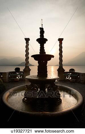 Ornamental gardens of Villa Monastero with carved stone wall, and spiral stone columns, with view overlooking Lake Como and mountain range beyond, sun setting across lake, Northern Italy View Large Photo Image Stock Image - Ornamental gardens of Villa Monastero with carved stone wall, and spiral stone columns, with view overlooking Lake Como and mountain range beyond, sun setting across lake, Northern Italy. Fotosearch