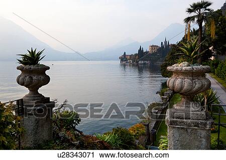 Stock Image - Ornamental gardens of Villa Monastero with carved stone wall, and terracotta pots on columns, with view overlooking Lake Como and mountain range beyond, autumn afternoon with blue sky. Fotosearch