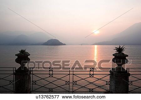 Ornamental gardens of Villa Monastero with ornate wrought iron railing, view overlooking Lake Como and mountain range beyond, sun setting across lake, Northern Italy View Large Photo Image Stock Photo - Ornamental gardens of Villa Monastero with ornate wrought iron railing, view overlooking Lake Como and mountain range beyond, sun setting across lake, Northern Italy. Fotosearch
