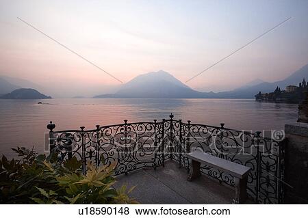 Stock Photo - Ornamental gardens of Villa Monastero with ornate wrought iron railing, stone bench on balcony, view overlooking Lake Como and mountain range beyond, sun setting across lake, Northern Italy. Fotosearch