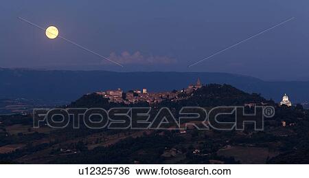 Stock Photograph - Panorama view of full moon rising over medieval hilltop town of Todi, with spire of San Fortunato, and Tempio of Bramante, town lights illuminated at dusk, Umbria, Italy. Fotosearch