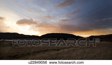 Stock Photograph - Panorama view of Lago Grey at sunrise, with dramatic orange and blue sky lighting scrub grass and distant hills silhouetted across lake, Torres del Paine, Southerm Chile, Patagonia. Fotosearch