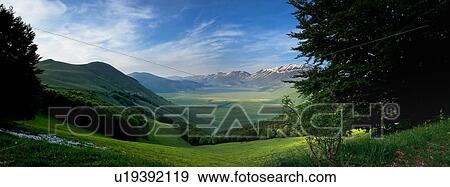 Panorama view of Piano Grande meadow, snow-capped mountains with hilltop town of Castelluccio in background, sunny spring evening, Umbria, Italy View Large Photo Image Stock Photo - Panorama view of Piano Grande meadow, snow-capped mountains with hilltop town of Castelluccio in background, sunny spring evening, Umbria, Italy. Fotosearch