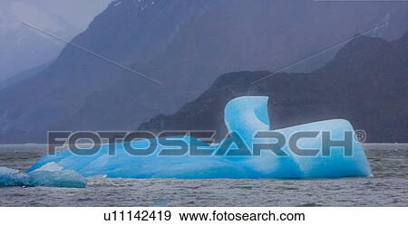Stock Photo - Panorama view of stormy grey Lago Grey with bright blue iceberg fragment floating in front of glacier, and snow covered mountains in background, Torres del Paine, Southern Chile, Patagonia. Fotosearch