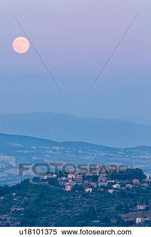 Stock Photography - Panoramic view of medieval hilltop town of Monte Castello di Vibio at sunset, hills and fields beyond, with full moon rising over village, pink light, and blue dusk sky, Umbria, Italy. Fotosearch