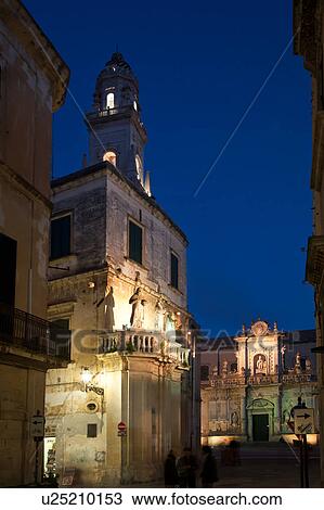 Stock Image - Piazza del Duomo, with view of campanile (bell tower) and Baroque ornamentation of Cathedral, dusk with night illumination, 3 figures in foreground, Lecce, Salento, Puglia, Southern Italy. Fotosearch
