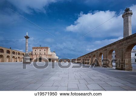Stock Image - Piazza surrounded by arched loggia, with Basilica, lighthouse and Corinthian column on sunny afternoon with stormy sky, Santa Maria di Leuca, Salento, Puglia, Southern Italy. Fotosearch