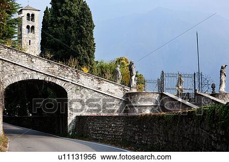 Stock Photograph - Renaissance stone arch over road, part of villa, with statues above and church tower in background, ivy growing along side stone wall on sunny autumn afternoon, Lake Como, Northern Italy. Fotosearch