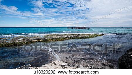 Stock Image - Rocky beach with bright blue water, red metal pier, cargo boat on horizon, and snow capped mountains of Albania visible across sea, dramatic clouds in blue sky, Salento, Puglia, Southern Italy. Fotosearch