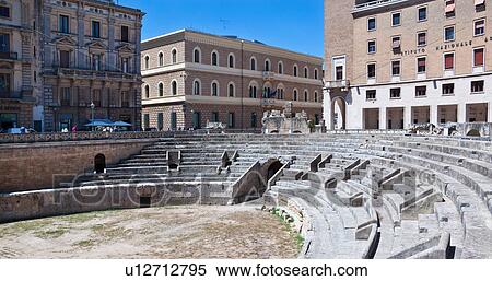 Stock Photography - Roman amphitheatre with semicircular arena in foreground; shops, Renaissance, Baroque and modern facades beyond, in morning sunshine with blue sky, Lecce, Salento, Puglia, Southern Italy. Fotosearch