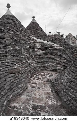 Stock Image - Rooftop view: town of Alberobello, World Heritage site, with trullo houses, white painted stone with piled stone conical roofs and large chimneys, monochrome view, Puglia, Southern Italy. Fotosearch