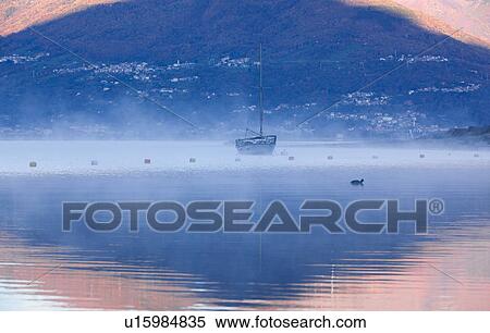 Stock Photography - Sailing boat moored at Lake Como,, golden light of sunrise reflected in water with swirling mists, backdrop of mountain slopes, Lombardy, Northern Italy.. Fotosearch
