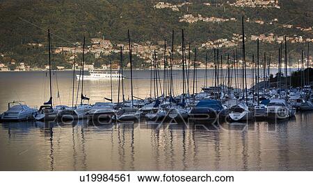 Stock Image - Sailing boats moored in harbour inlet, Lake Como, in early morning golden light, masts reflected in calm water, with passenger ferry and mountain slope of opposite shore beyond, Lake Como, Northern Italy. Fotosearch