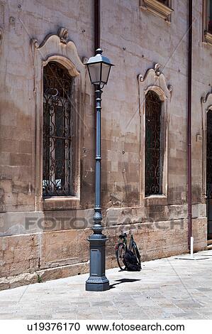 Stock Image - Sreet view detail with facade of Renaissance palazzo, wrought iron grille over 2 windows, metal lamp post, and parked bicycle against palace wall, Lecce, Salento, Puglia, Southern Italy. Fotosearch