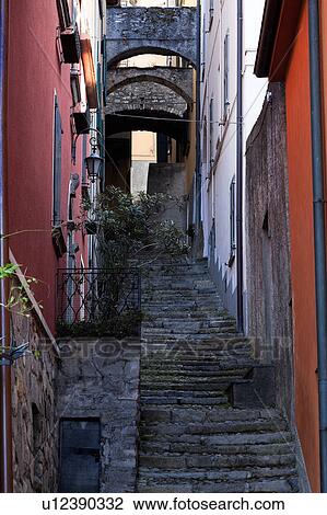 Stock Image - Steep uneven stone stairway up narrow alley with walls of houses painted red/orange in typical evocative style of Northern Italy. Fotosearch