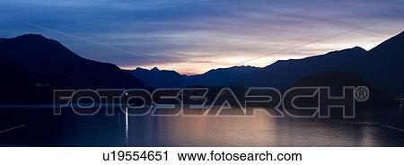 Stock Image - Stitched panorama view across Lake Como at dusk, with beautiful calm sunset colours of blue, purple and pastel pink, mountains silhouetted beyond Bellagio, with town lights illuminated, Lake Como, Northern Italy. Fotosearch