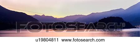 Stock Image - Stitched panorama view across Lake Como at dusk, with beautiful calm sunset colours of purple and pastel pink, mountains silhouetted beyond Bellagio, with town lights illuminated, Lake Como, Northern Italy. Fotosearch