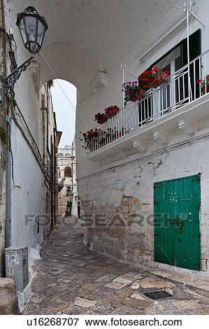 Stock Photo - Street in Ostuni, with typical white stucco walls, stone paving, wrought iron lamp, and archway, with flowers decorating, spring afternoon, Salento, Puglia, Southern Italy. Fotosearch